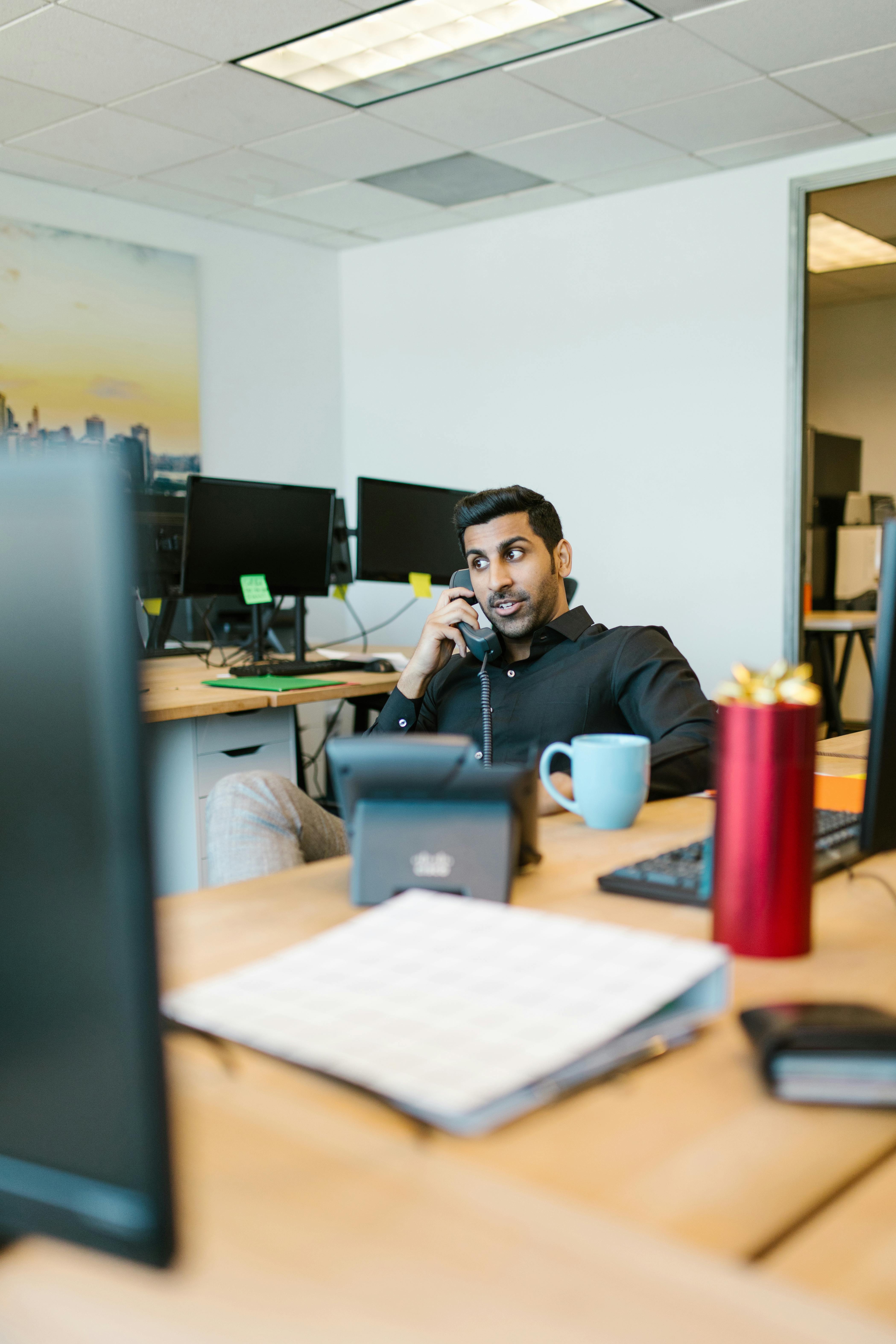 A focused young professional talking on the phone in a modern office setting, surrounded by computers.